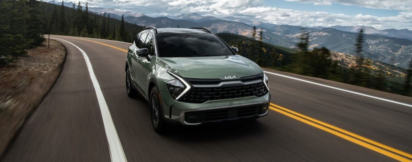 A green 2023 Kia Sportage driving a paved road with mountains in the background.